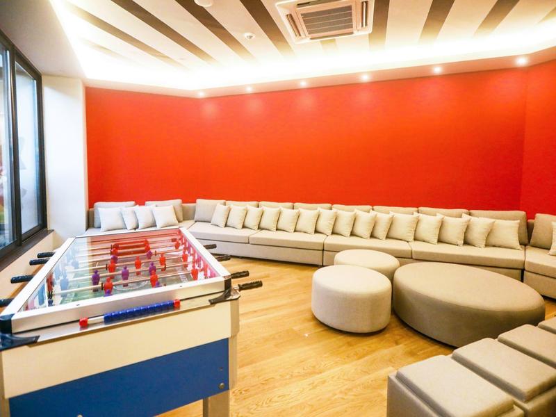 Lounge luminosa con parete rossa, angolo divano grigio lungo, sgabelli e calcio balilla