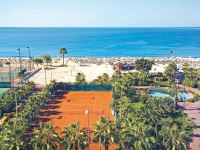 View of tennis courts, palm trees, and a beach with blue sea in the background.