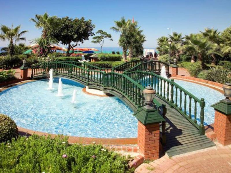 Green pedestrian bridge over small pool with fountains and palm trees at vacation spot.