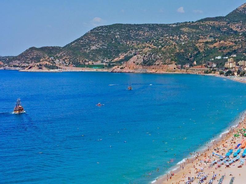 Long sandy beach with umbrellas and clear blue sea, mountains in the background.