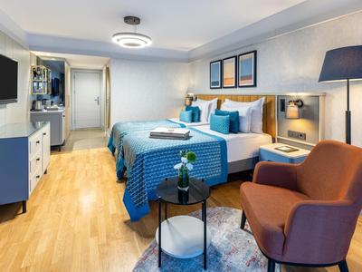 Moderne hotelkamer met twee bedden, blauwe inrichting en zithoek met fauteuil en lamp.