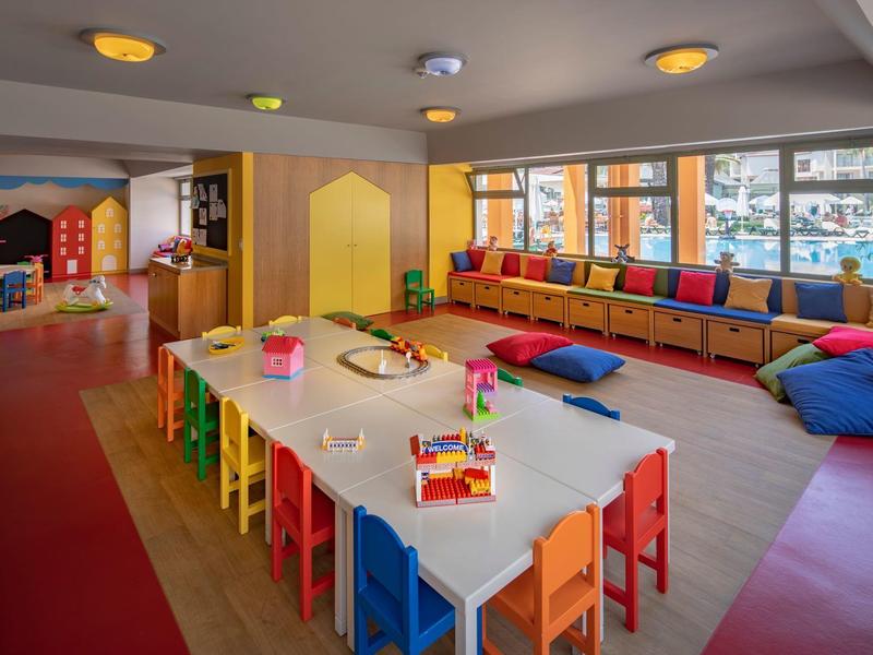 Sala giochi colorata per bambini con tavoli, sedie e cuscini vicino a grandi finestre.