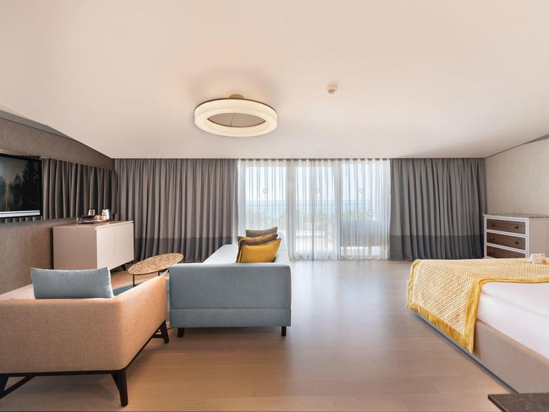 Camera d'hotel moderna con zona salotto, letto grande e finestre dal pavimento al soffitto con tende.