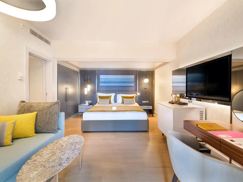 Camera d'hotel luminosa con letto grande, TV moderna, scrivania e area salotto accogliente.