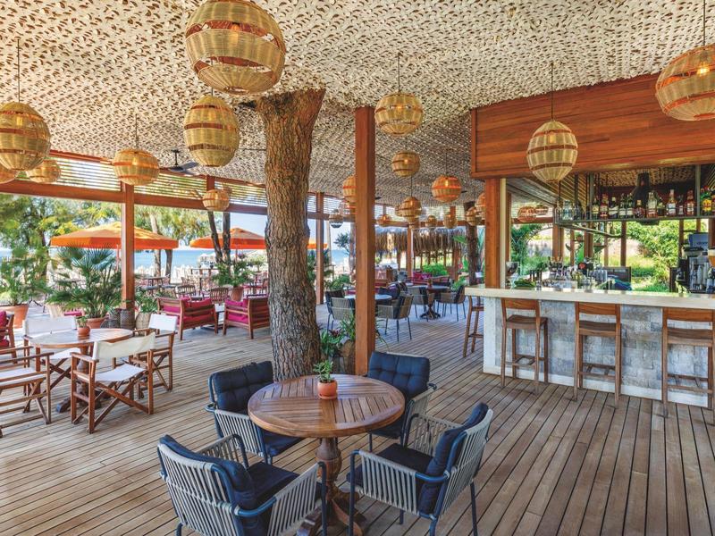 Bar all'aperto con pavimento in legno, tavoli rotondi e sedie confortevoli in ambiente tropicale.