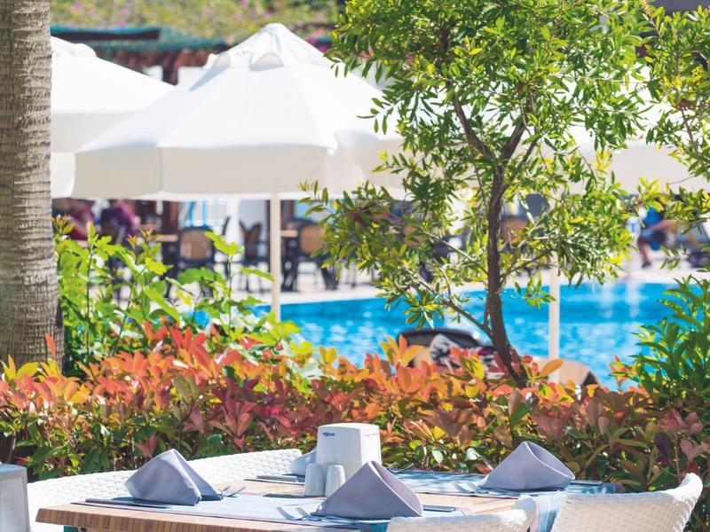Outdoor poolside dining area with white chairs, tables, umbrellas, and green foliage.