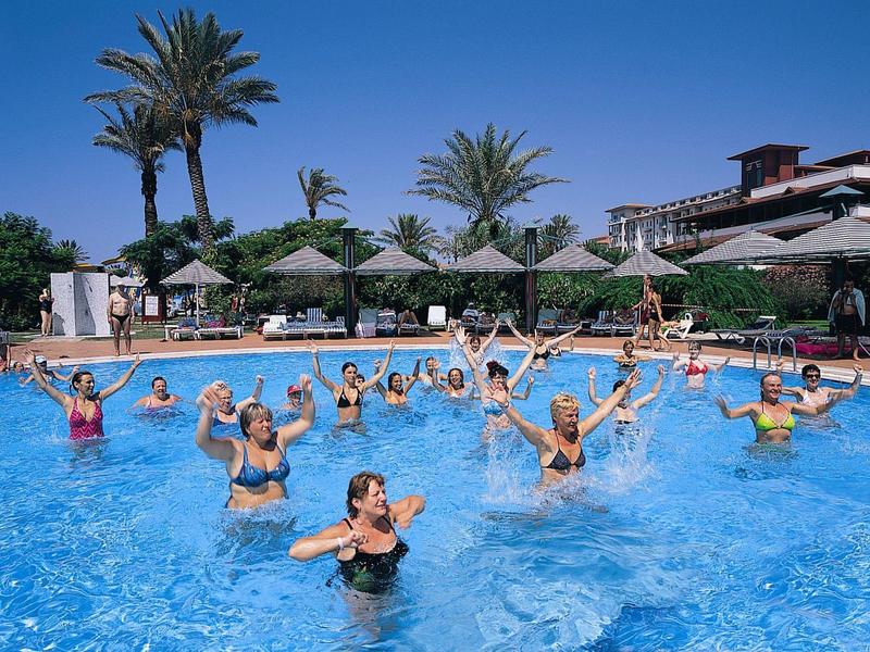 Grupa osób na hotelowym basenie podczas aqua aerobiku pod błękitnym niebem z palmami.