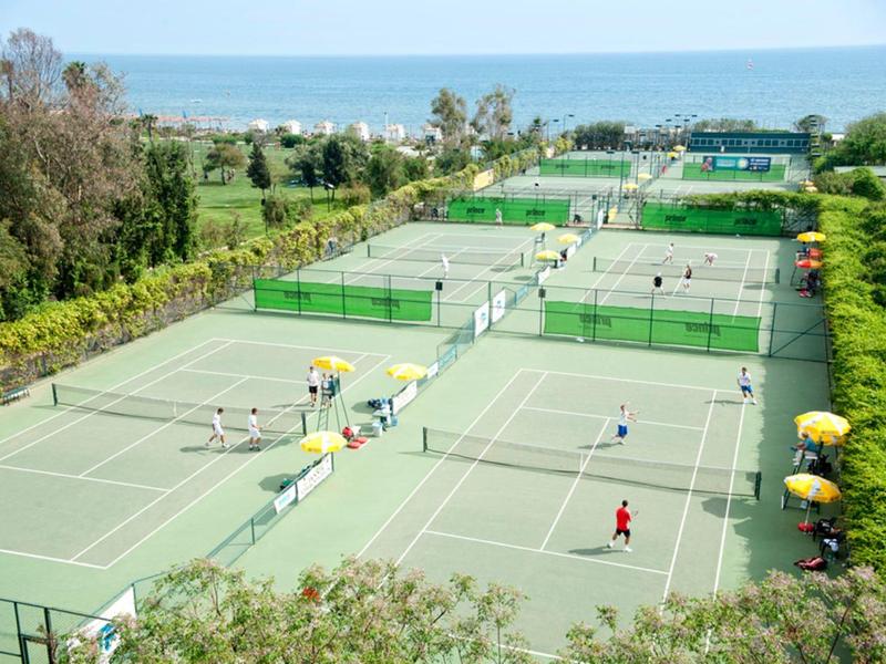 Kilka kortów tenisowych z zawodnikami i żółtymi parasolami, morze w tle.