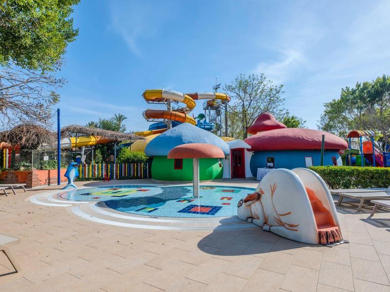 Kolorowy park wodny hotelu z zjeżdżalniami i fontanną w kształcie grzyba pod niebieskim niebem.