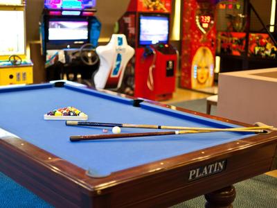 Billardtisch mit blauem Filz, Billardkugeln, zwei Queues und Arcade-Spielen im Hintergrund.