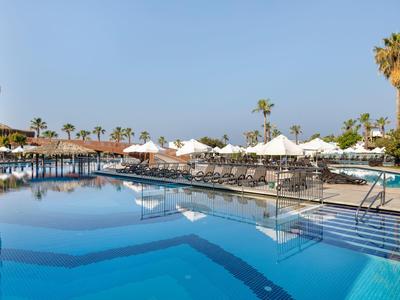 Großer Pool mit klar blauem Wasser, Liegen und Schirmen, Palmen und klarem Himmel im Hintergrund.