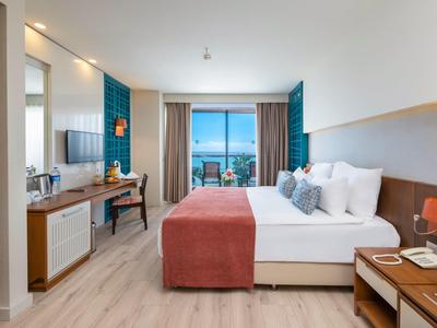 Helles Hotelzimmer mit Doppelbett, Schreibtisch, Fernseher und Balkon mit Meerblick.