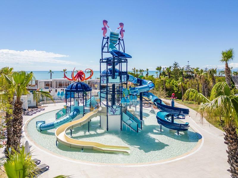 Parco acquatico con scivoli d'acqua e palme, cielo azzurro e mare sullo sfondo.
