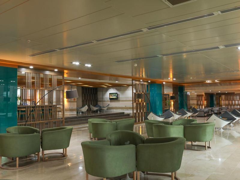 Sala d'attesa dell'aeroporto con sedie verdi e area d'imbarco luminosa.