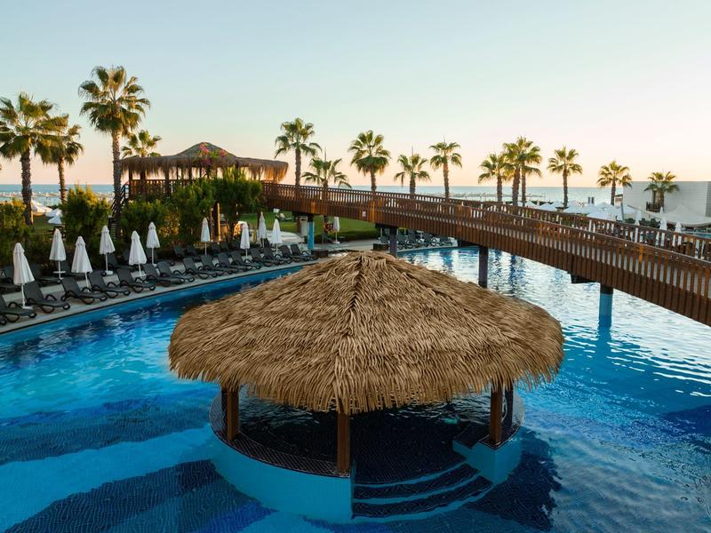 Piscina con bar palafitta, palme e passerella in legno al tramonto.