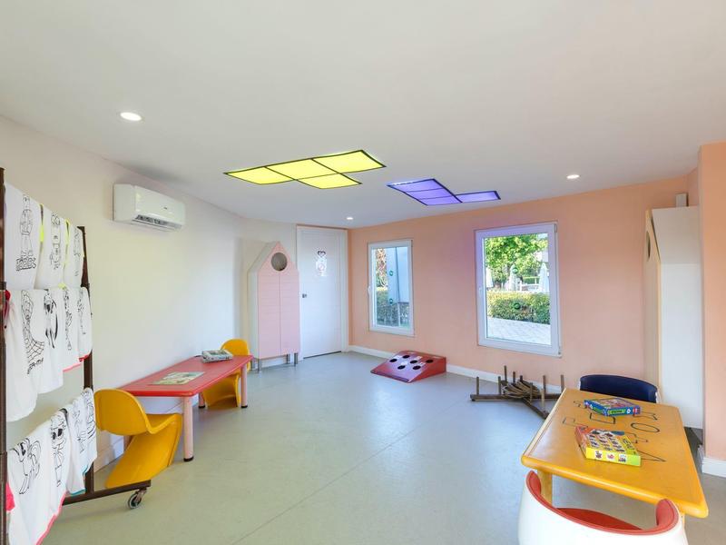 Sala giochi colorata e luminosa con tavoli, sedie e angolo lettura per bambini.