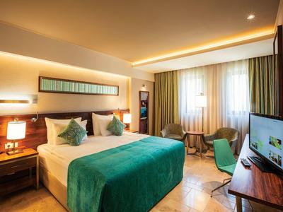Modern eingerichtetes Hotelzimmer mit Doppelbett, grünem Bettüberwurf, Schreibtisch und TV.