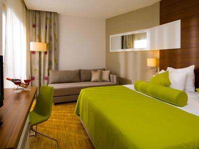 Modernes Hotelzimmer mit grünem Bett, Sofa, Fernseher und bodentiefen Fenstern.