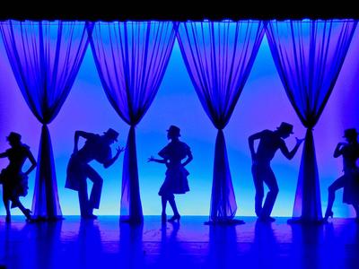 Blaue Bühne mit fünf tanzenden Silhouetten vor leuchtenden Vorhängen.