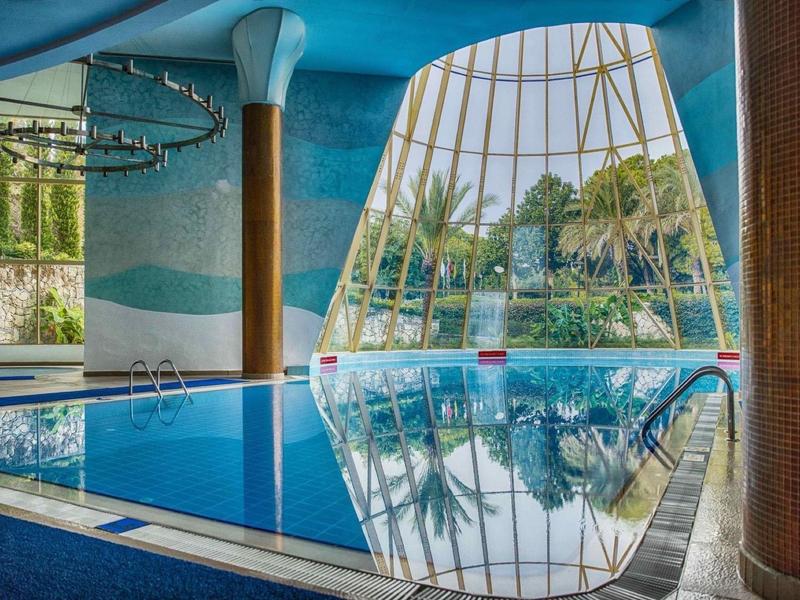 Innenraum eines Hallenbades mit großem Schwimmbecken, Glasdach und Palmen im Hintergrund.