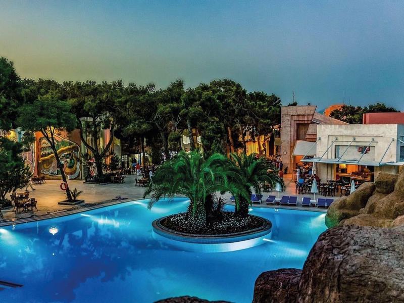 Großer beleuchteter Pool mit Insel und Palmen, umgeben von Bäumen und Gebäuden bei Dämmerung.