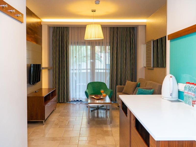 Helles Wohnzimmer mit Küchentheke, grünem Sessel, Sofa, großem Fenster und grauen Vorhängen.