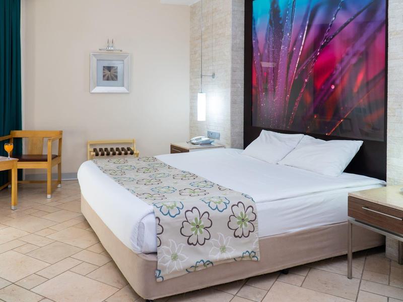 Modernes Schlafzimmer mit großem Bett, buntem Wandbild, Holzmöbeln und hellem Fliesenboden.