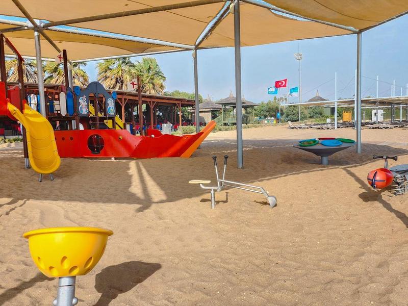 Kinderspielplatz mit Sand, buntem Piratenschiff, Schaukeln und Sonnensegel unter blauem Himmel.