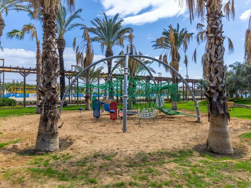 Kinderspielplatz mit Klettergerüst, Sandfläche und Palmen unter blauem Himmel und Wolken.