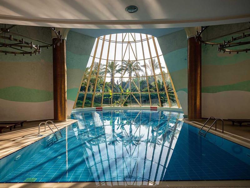 Hallenbad mit großem, kegelförmigem Fenster und Liegestühlen, das Poolwasser spiegelt die Decke.