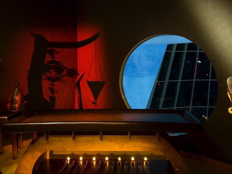 Dunkler Raum mit rötlicher Beleuchtung, runder blauer Fensteröffnung und goldener Büste auf Sockel.