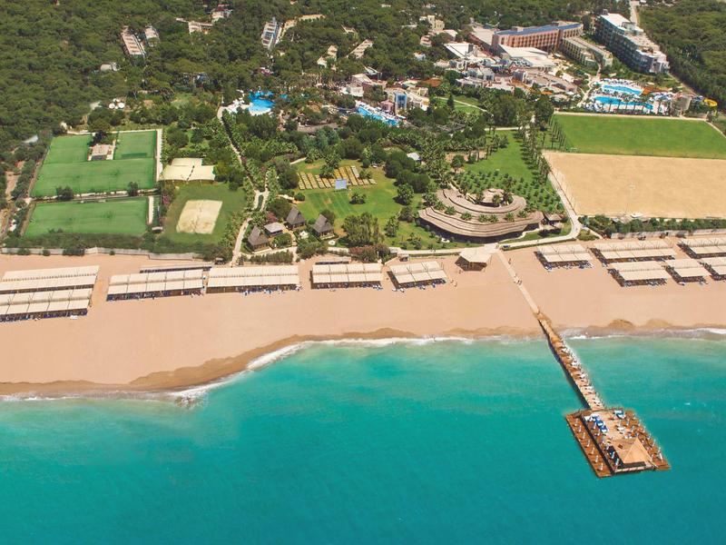 Luftaufnahme eines Hotelresorts mit Pool, Tennisplätzen, Strand und Steg ins türkisfarbene Meer.