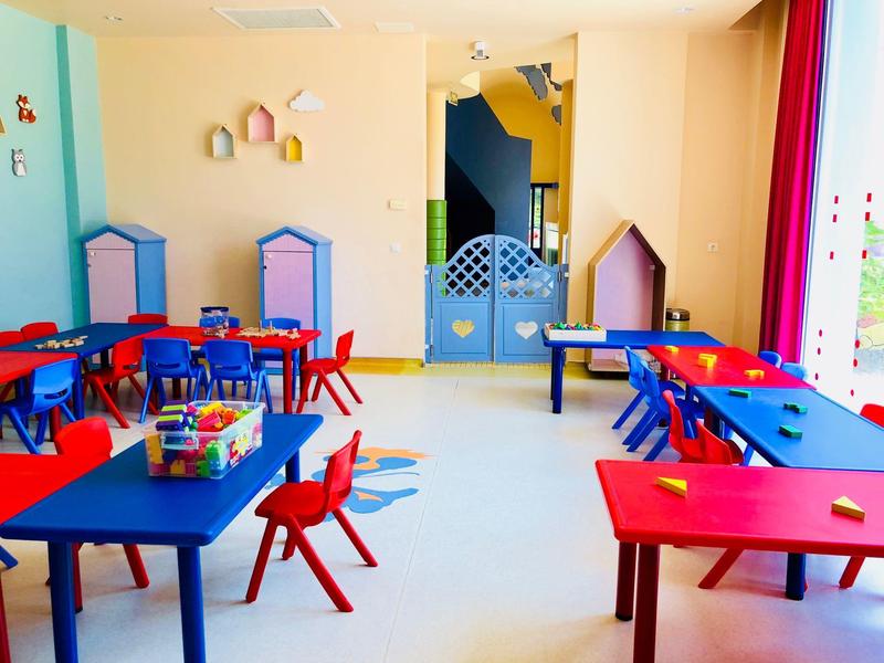 Buntes Klassenzimmer mit kleinen Tischen, roten Stühlen und Spielzeug an den Wänden.