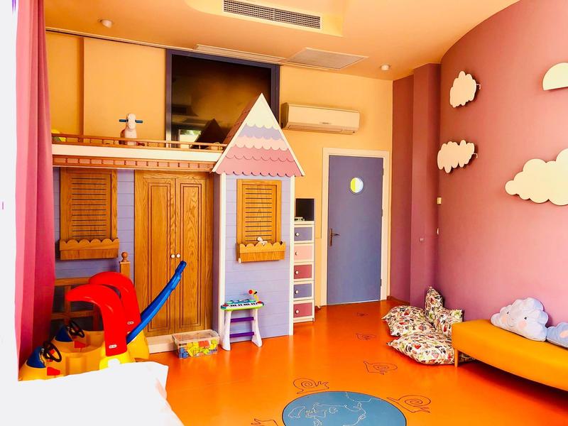 Buntes Kinderzimmer mit Spielhaus, Sofa, Wolken an der Wand und buntem Spielzeug auf orangem Boden.