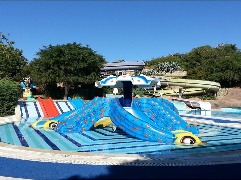 Wasserrutsche mit blauer Schlange in einem Becken, umgeben von Bäumen und blauem Himmel.