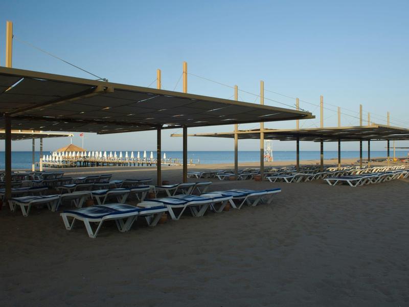 Leere Sonnenliegen unter großen Sonnendächern am Strand mit ruhigem Meer im Hintergrund.
