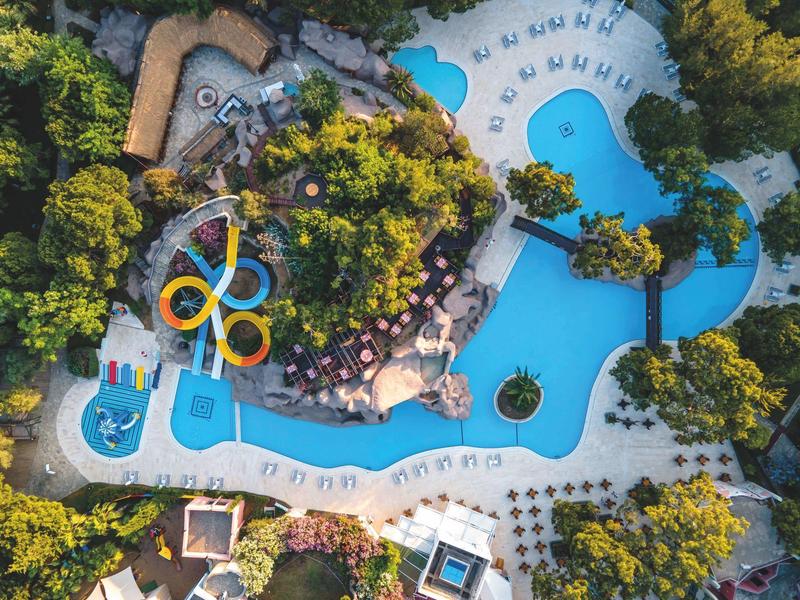 Luftaufnahme eines Wasserparks mit Pools, Rutschen und vielen Sonnenliegen, umgeben von Bäumen.
