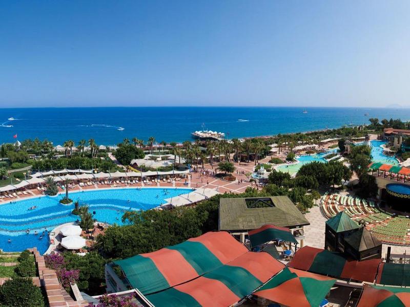 Scenic resort view with large swimming pools, colorful umbrellas, and blue sea on the horizon.