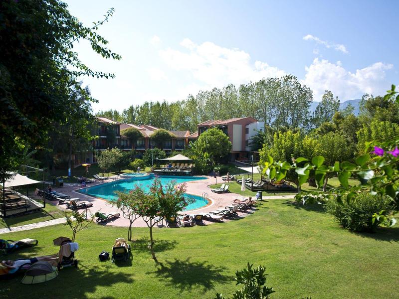 Sunny outdoor hotel pool surrounded by green lawn, trees, and lounge chairs with guests relaxing.