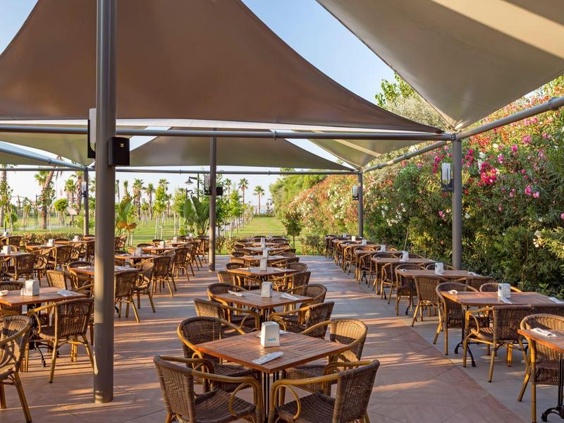 Outdoor hotel restaurant with wooden tables and chairs under a canopy surrounded by greenery.