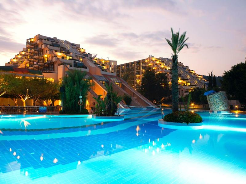 Lit resort pool at dusk with modern buildings and palm trees in the background
