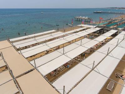 Vogelperspektive auf einen Strand mit weißen Sonnensegeln, Liegestühlen und blauem Meer im Hintergrund.