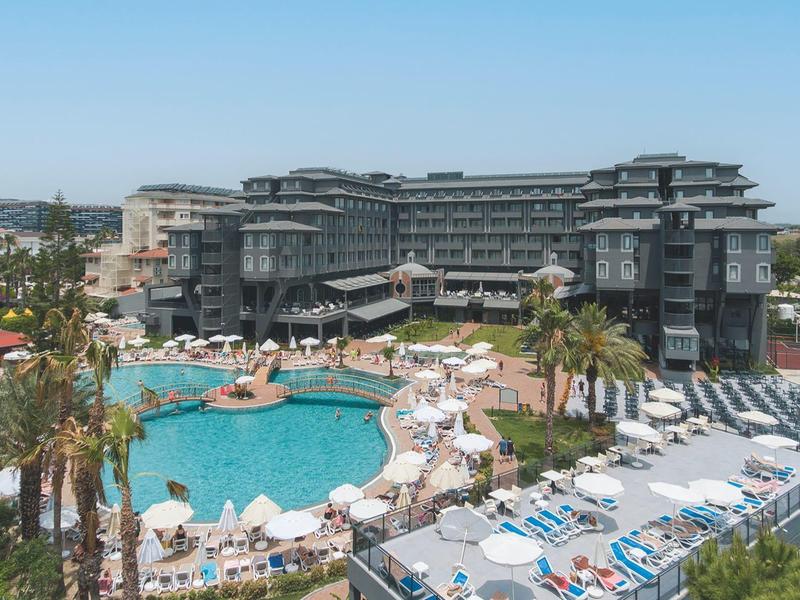 Complesso alberghiero moderno con grande piscina, ombrelloni e palme sotto un cielo limpido.