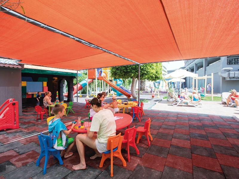 Bambini che giocano e si siedono sotto una vela arancione in un parco giochi.