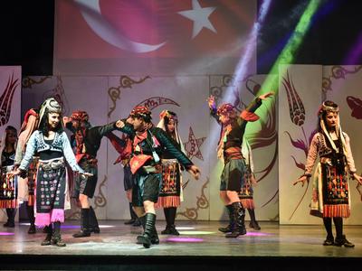 Menschen tanzen in bunter, traditioneller Tracht auf einer Bühne mit türkischer Flagge im Hintergrund.