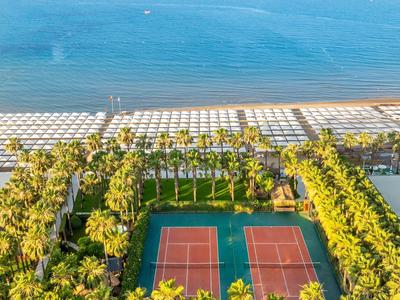 Strand mit weißen Sonnenschirmen, dahinter Tennisplätze und Palmen an der Küste.