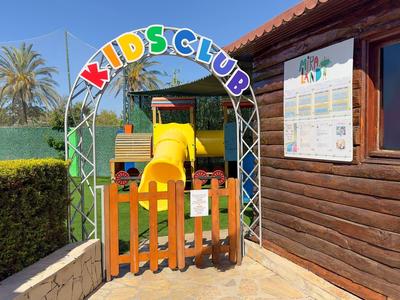 Bunter Eingang mit Gitter zum Kidsclub, gelbe Rutsche sichtbar, neben Holzwand mit Fenster und Kalender.