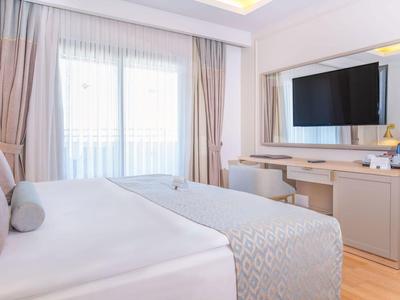 Helles Schlafzimmer mit Doppelbett, großem Spiegel, Fernseher und Fenster mit Vorhängen.