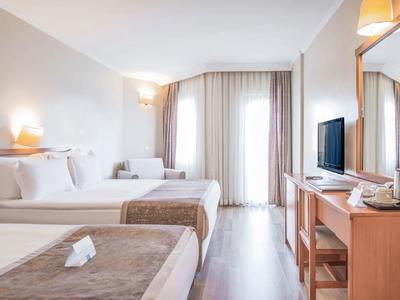 Helles Hotelzimmer mit zwei Betten, Holzmöbeln, TV und großen Fenstern mit Vorhängen.