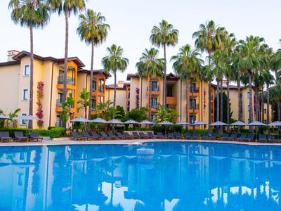 Grande piscina davanti a un hotel con palme e ombrelloni sotto un cielo sereno.